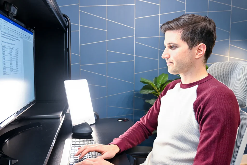 a young guy sitting in front of his computer using a happy lamp