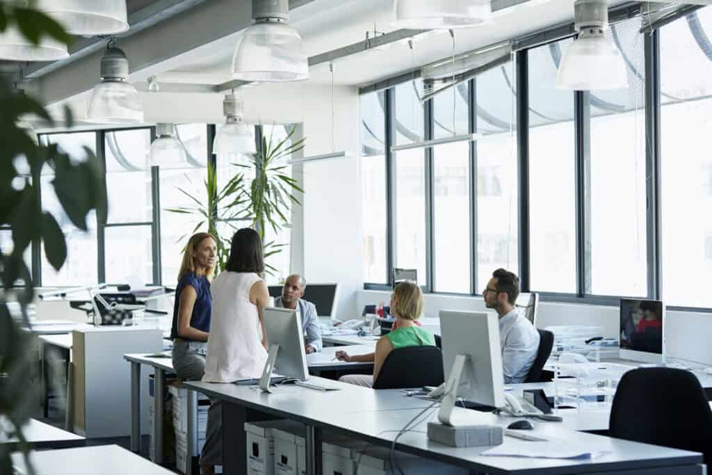 Bright modern office with large windows allowing natural light, where a group of professionals are gathered at desks for discussion, illustrating the importance of view direction for melanopic light exposure.