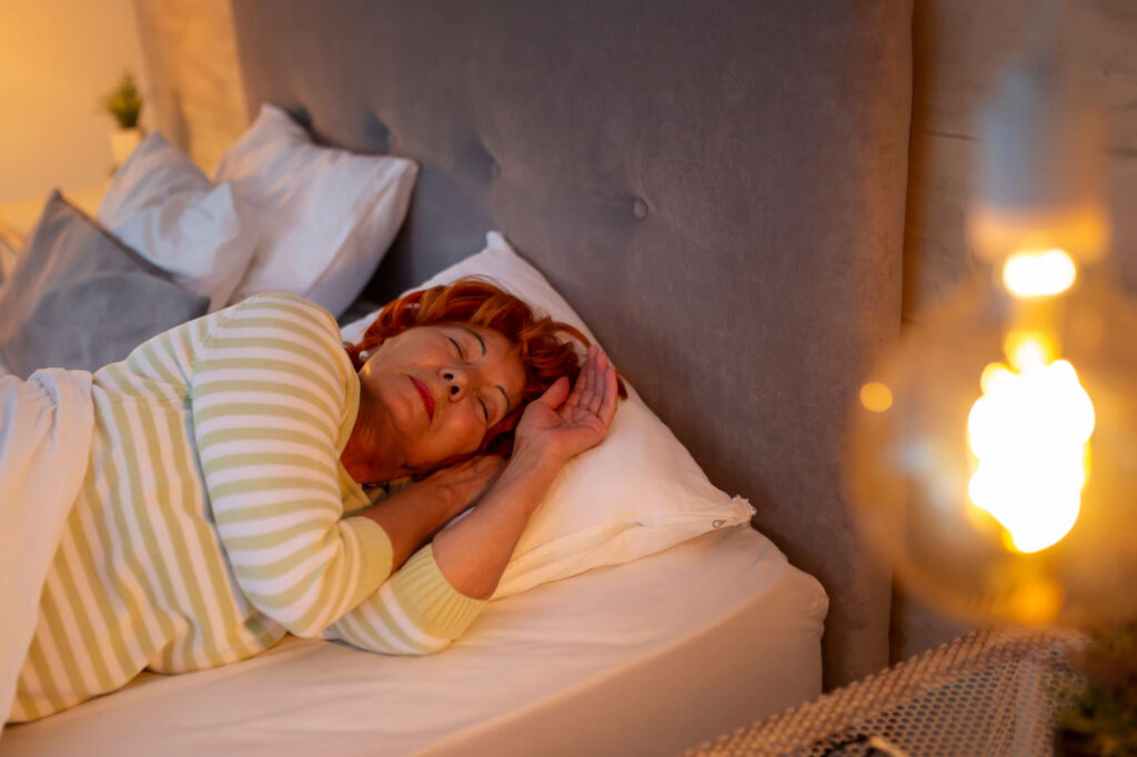 An elderly woman asleep in her bed. There is an incandescent light in the foreground.